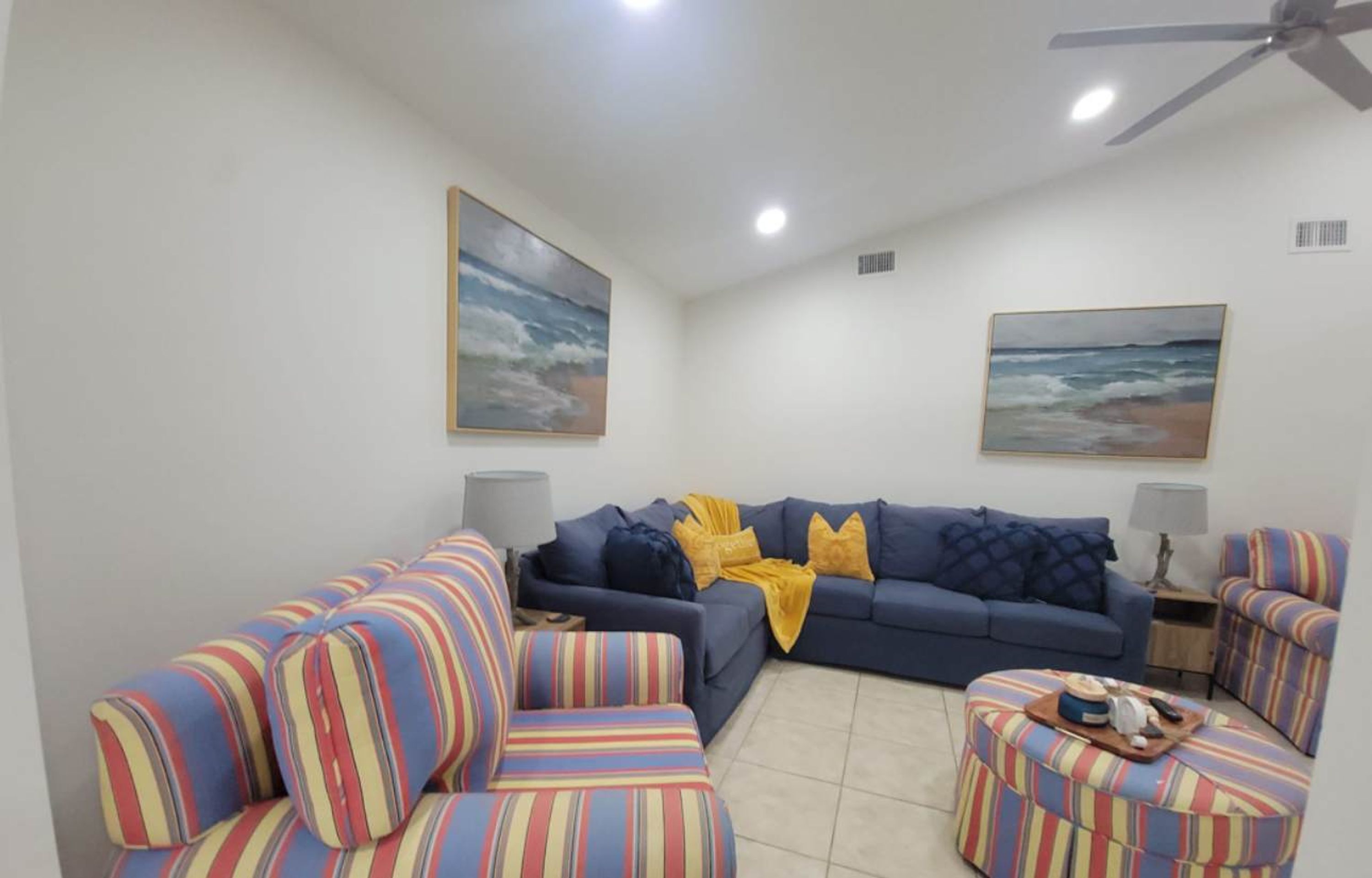 A living room with a blue sofa, striped armchairs, and ottoman, a ceiling fan, and two paintings on the walls.