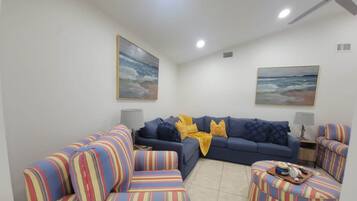 A living room with a blue sofa, striped armchairs, and ottoman, a ceiling fan, and two paintings on the walls.