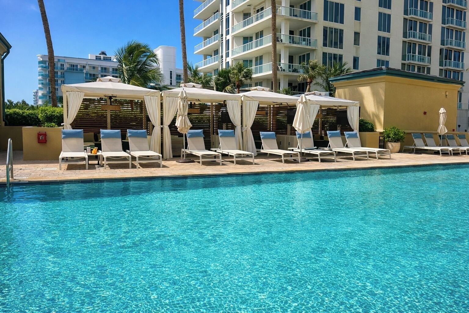 A swimming pool with lounge chairs and umbrellas, surrounded by palm trees and a multi-story building in the background.