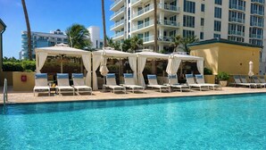Una piscina con sillones de playa y sombrillas, rodeada de palmeras y un edificio de varios pisos al fondo.