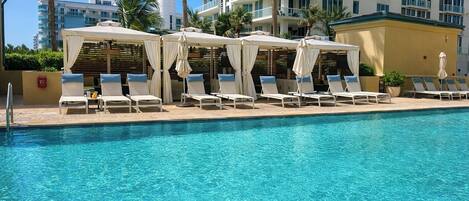 A swimming pool with lounge chairs and umbrellas, surrounded by palm trees and a multi-story building in the background.