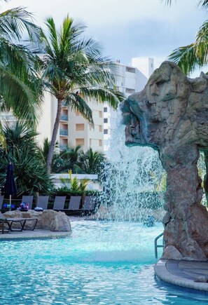 Ein Pool mit Wasserspiel, Palmen und einem Gebäude im Hintergrund.