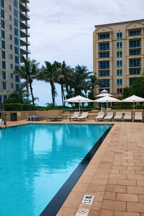 A swimming pool with lounge chairs and umbrellas, surrounded by palm trees and a multi-story building. - Ocean view! 2bd at The Palm Beach Resort and Spa (Riviera Beach)