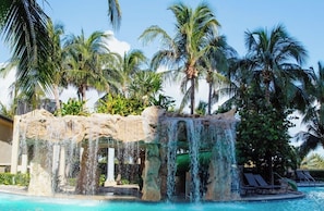 Uma área de piscina com cachoeira, palmeiras e espreguiçadeiras.