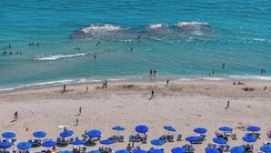Una platja amb persones nedant i prenent el sol, i diverses ombrelles blaves.
