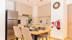 A modern kitchen with a stainless steel refrigerator, wooden dining table, and chairs. There is a fire extinguisher mounted on the wall and a clock above the sink. - Bloomfields Stylish 1BR in Waters Edge (Abu Dhabi)