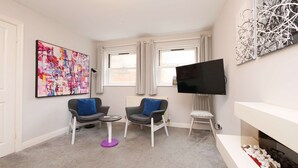 Un salon moderne doté d’un téléviseur à écran plat, de deux fauteuils gris avec des coussins bleus, d’une petite table ronde et d’une cheminée.