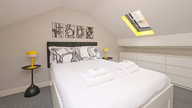 Un lit bien fait, avec du linge de lit et des serviettes blanches, une lampe moderne, des œuvres d’art abstraites accrochées au mur et un design de plafond original.