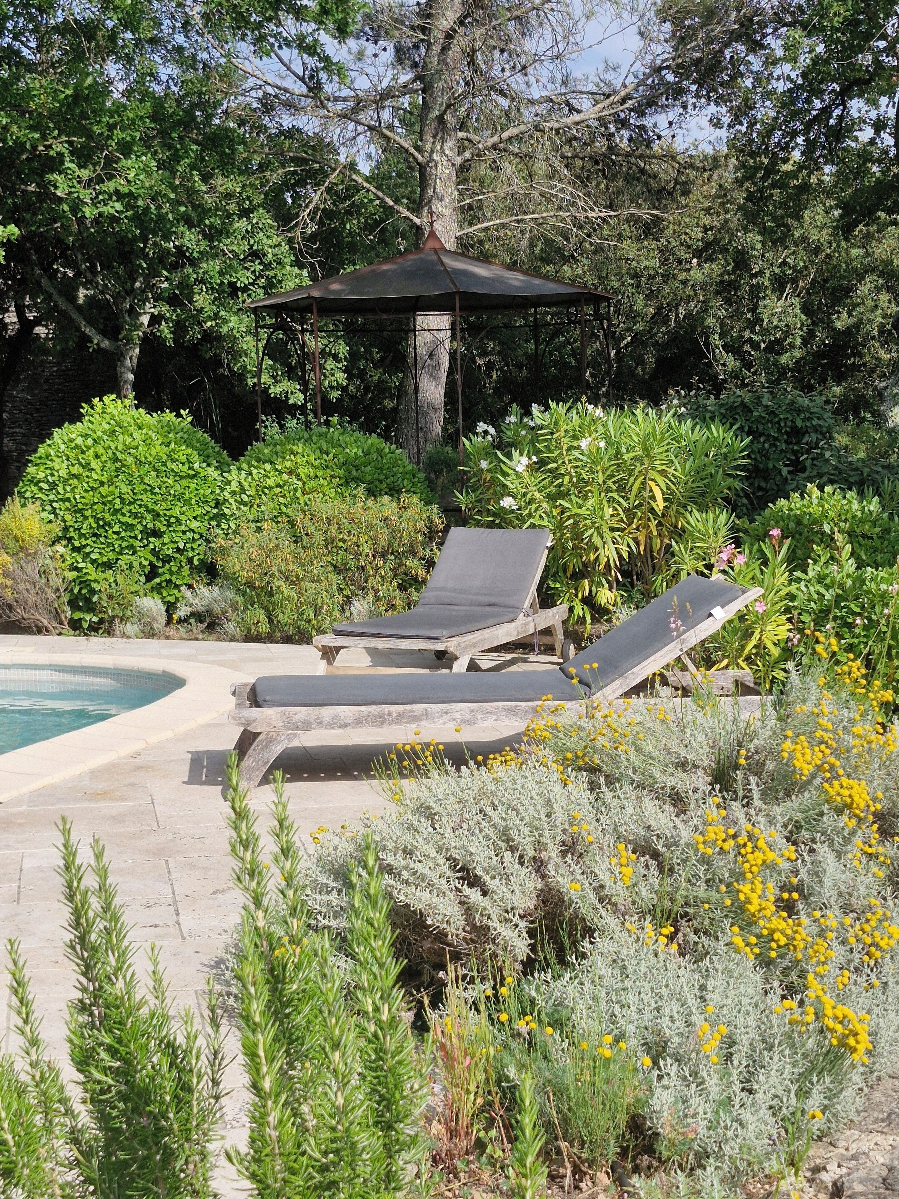 A pool area with lounge chairs and a gazebo surrounded by lush greenery and flowering plants.