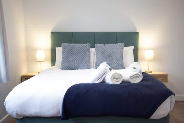Una cama bien hecha con una colcha blanca y azul, dos almohadas y toallas enrolladas sobre una falda de cama azul oscuro. Hay dos mesitas de noche con lámparas.