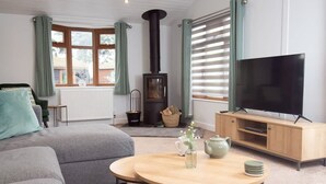 Ein gemütliches Wohnzimmer mit einer Couch, einem runden Holztisch mit einer Vase und Teekanne, einem Fernseher auf einer Holzkommode und einem schwarzen Kaminofen.