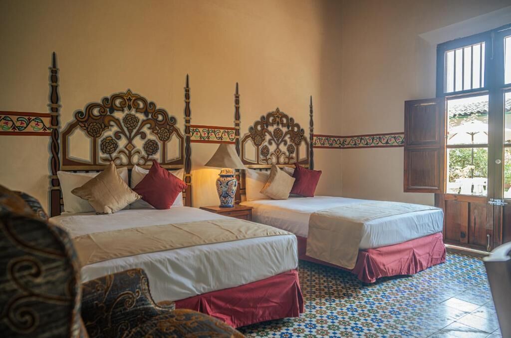 A hotel room with two beds, ornate headboards, a tiled floor, and a view through an open door.