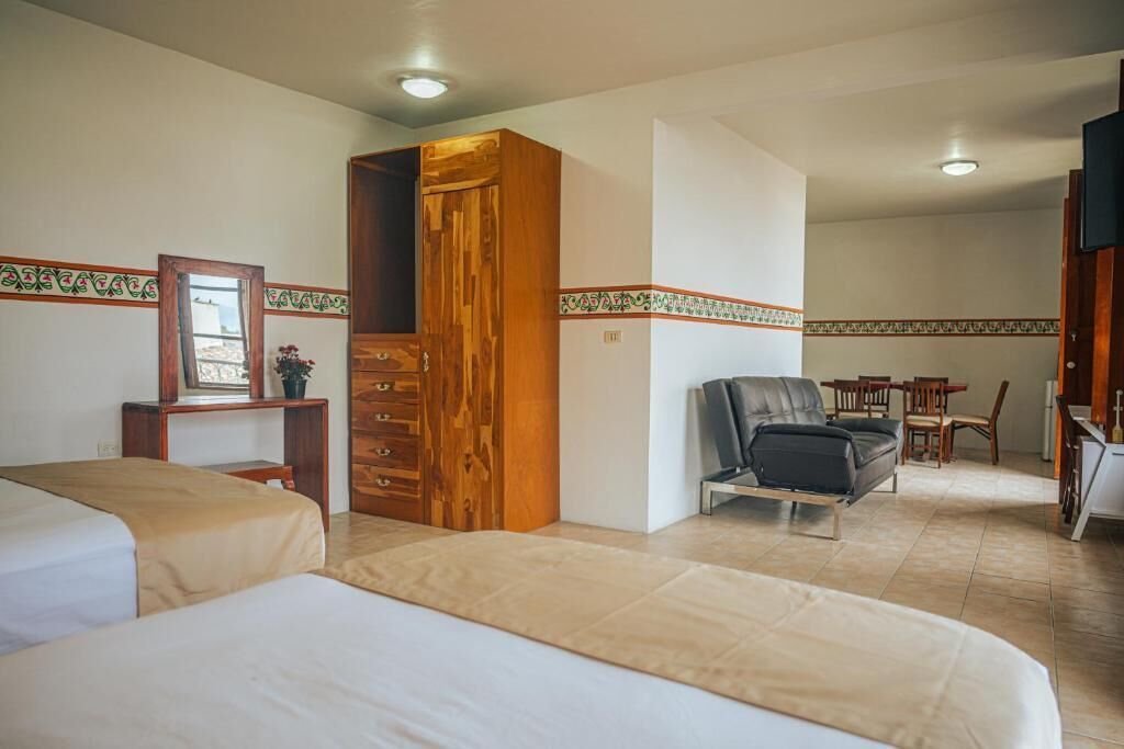 A hotel room with two beds, a wooden wardrobe, a desk with a chair, a sofa, and a dining area with a table and chairs.