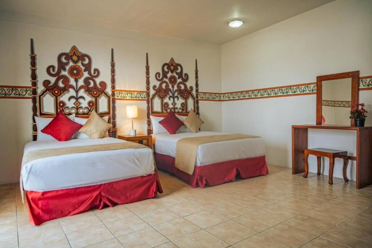 Ein Hotelzimmer mit zwei Betten, jeweils mit einem Holz-Kopfschild, einem kleinen Nachttisch mit Lampe und einem Holztischchen mit einer Vase.