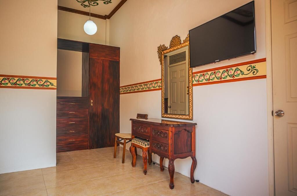 Una habitación con un armario de madera, una pequeña silla de madera, un espejo y un televisor colocado en la pared.