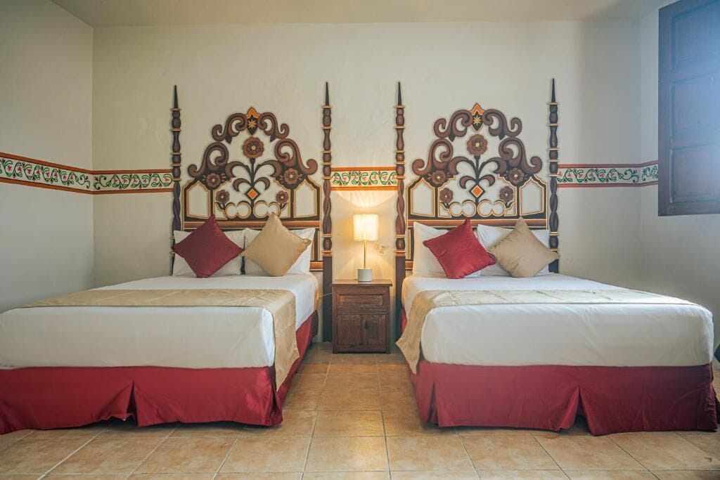 A hotel room with two beds, each with a decorative headboard, a nightstand with a lamp, and a tiled floor.