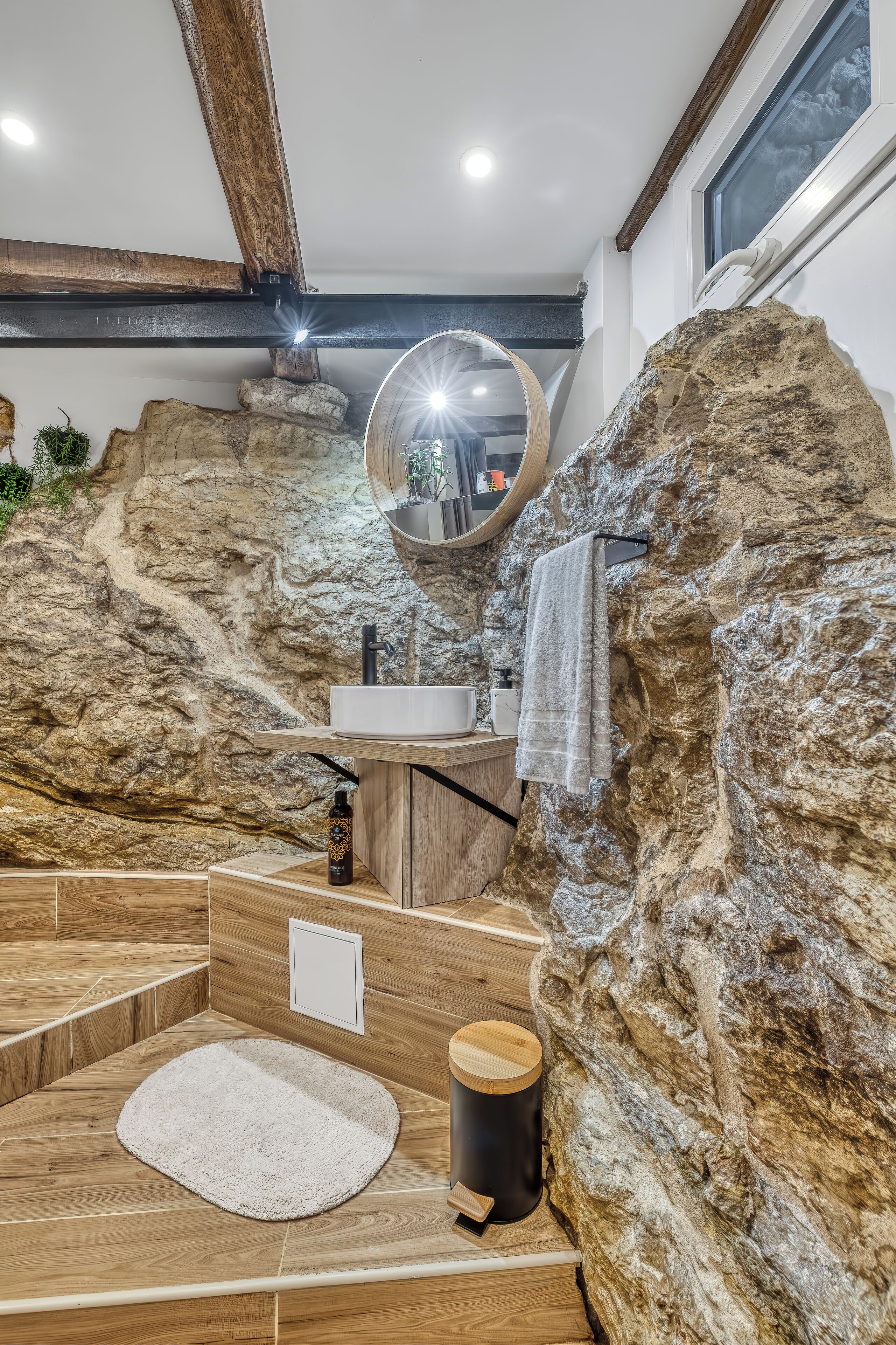 A bathroom with a rustic design, featuring a stone wall, wooden flooring, a round mirror, a sink, and a towel rack.