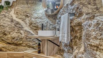 A bathroom with a rustic design, featuring a stone wall, wooden flooring, a round mirror, a sink, and a towel rack.