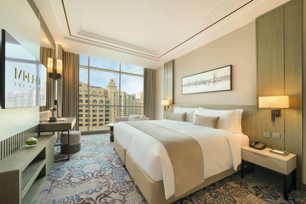 A hotel room with a large bed, a desk, a chair, a TV, and a window with a city view.