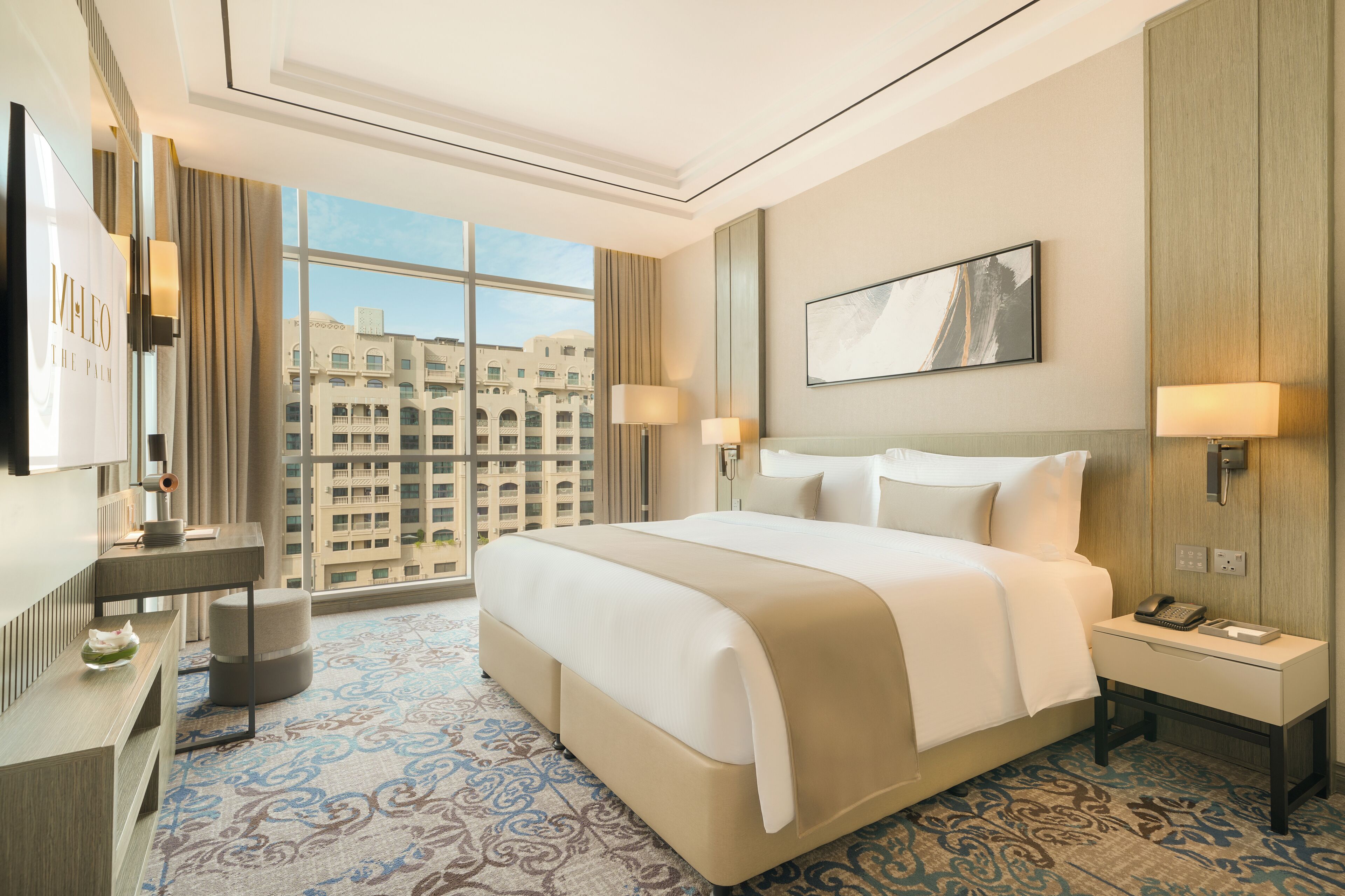 Ein Hotelzimmer mit einem großen Bett, Nachttischlampen, einem Schreibtisch und Blick auf die Stadt durch große Fenster.
