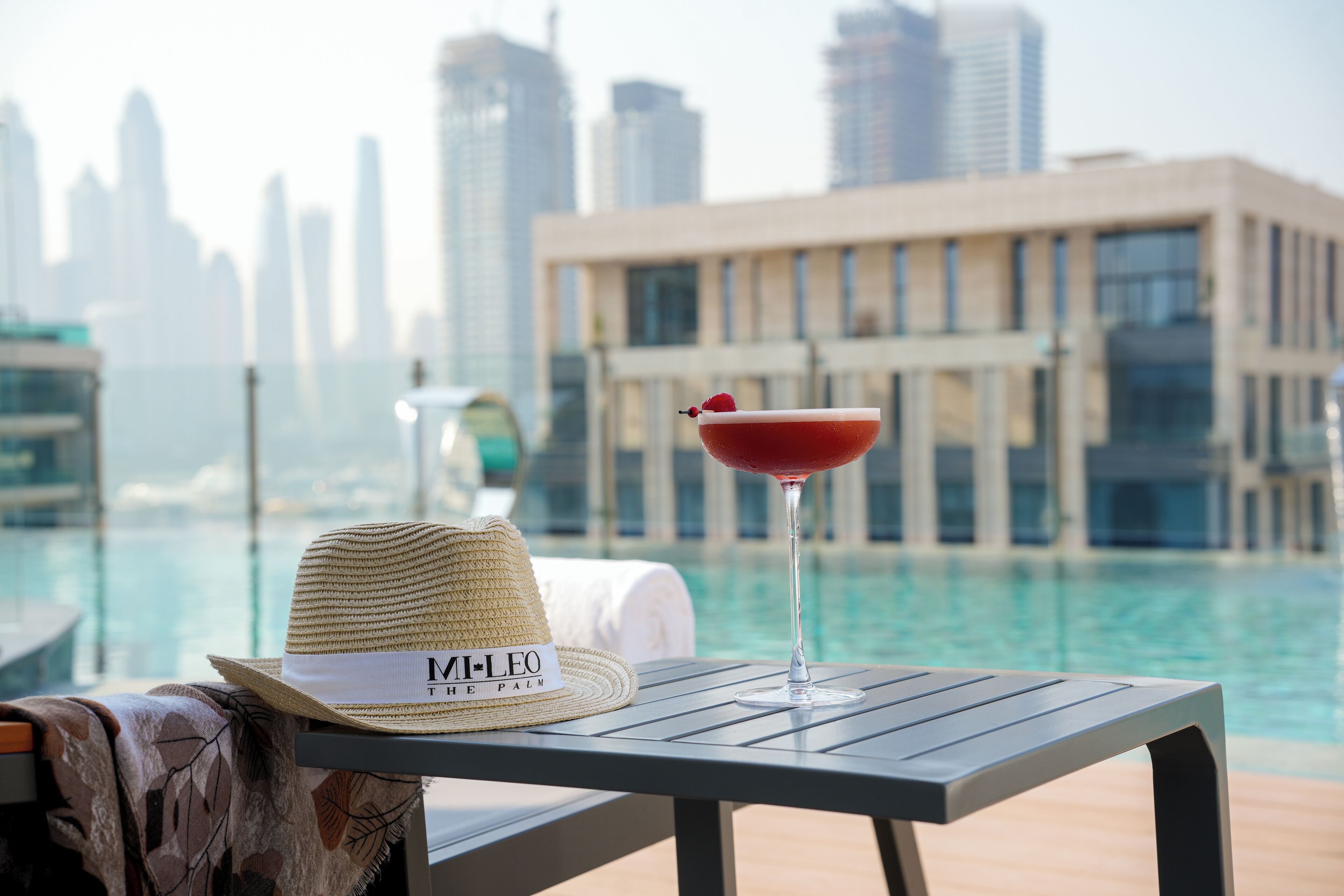 Un sombrero y un cóctel sobre una mesa junto a una piscina, con un horizonte urbano al fondo.