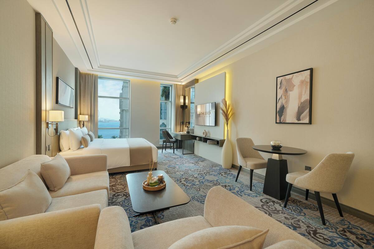 A hotel room with a bed, two sofas, a coffee table, a desk with a chair, a TV, and a large window offering a view.