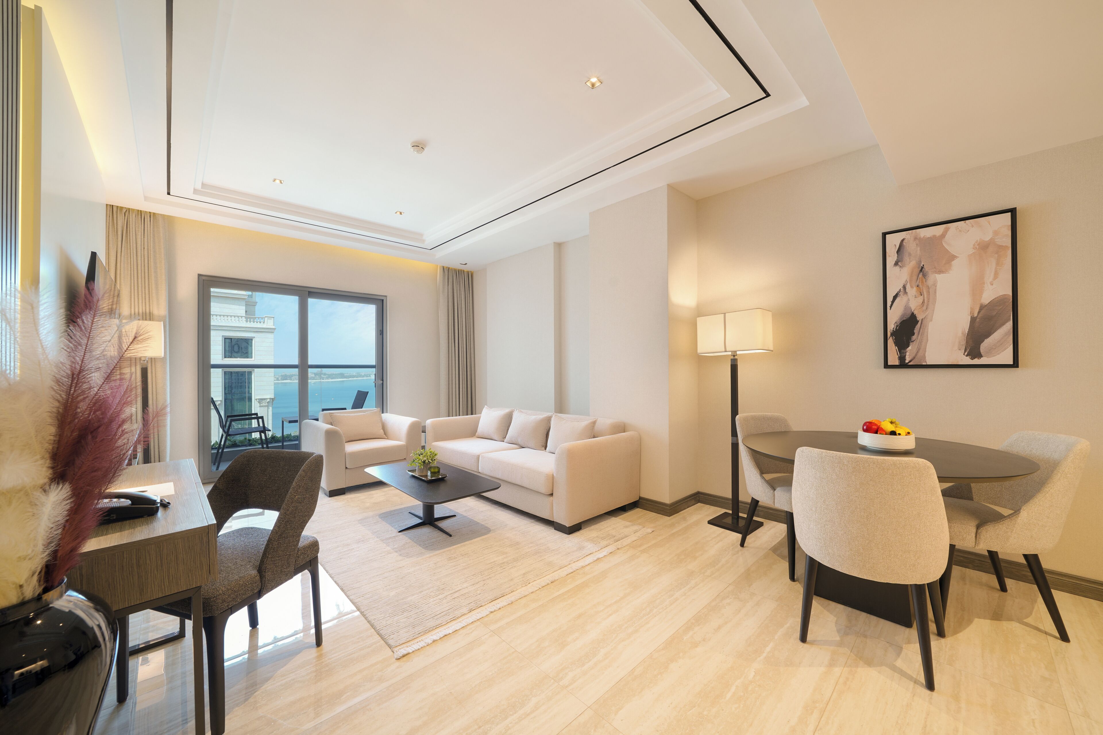 A modern hotel room with a sofa, dining table, chairs, a lamp, and a painting on the wall.