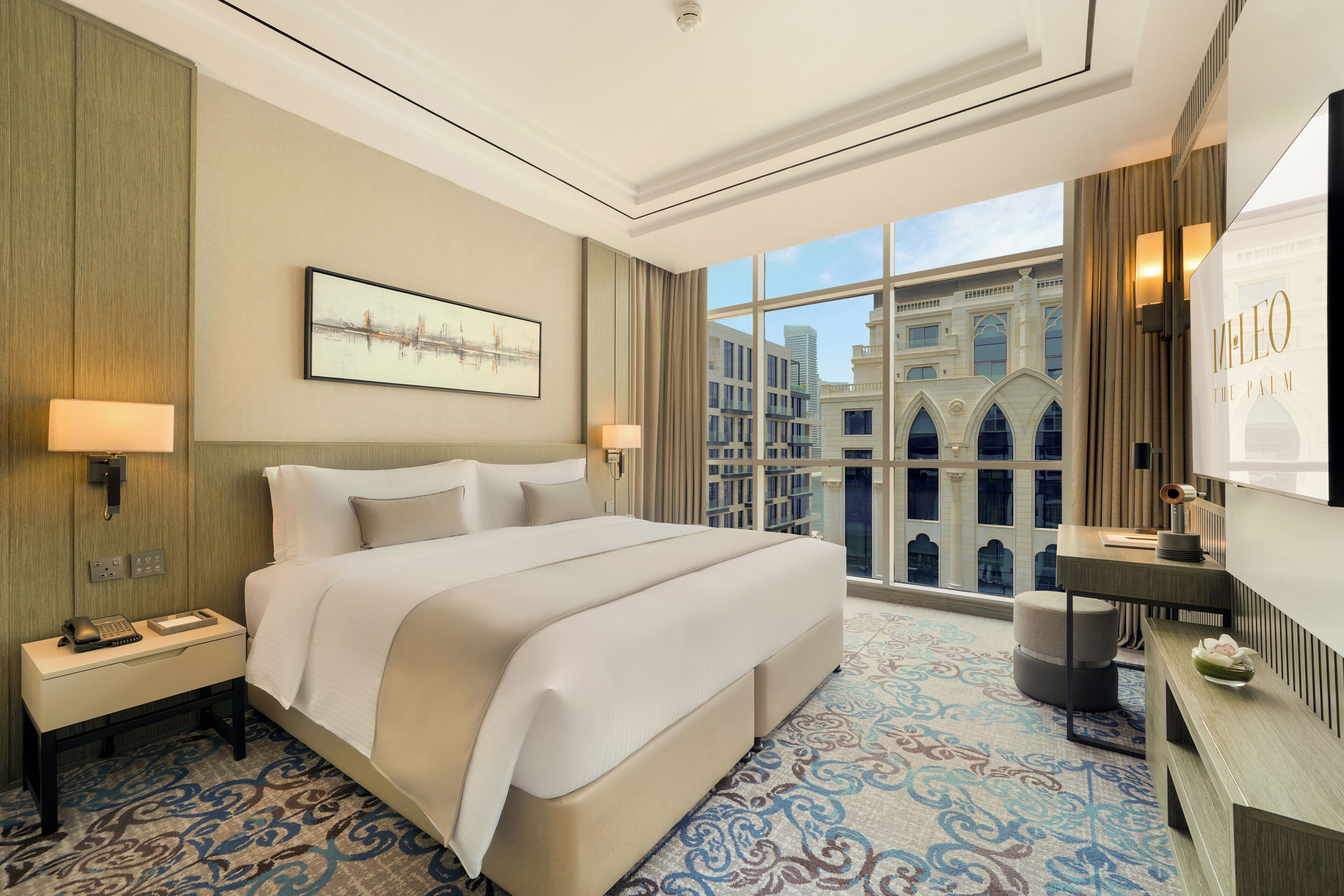 A hotel room with a large bed, a desk, a chair, a lamp, a telephone, and a view of a building through the window.