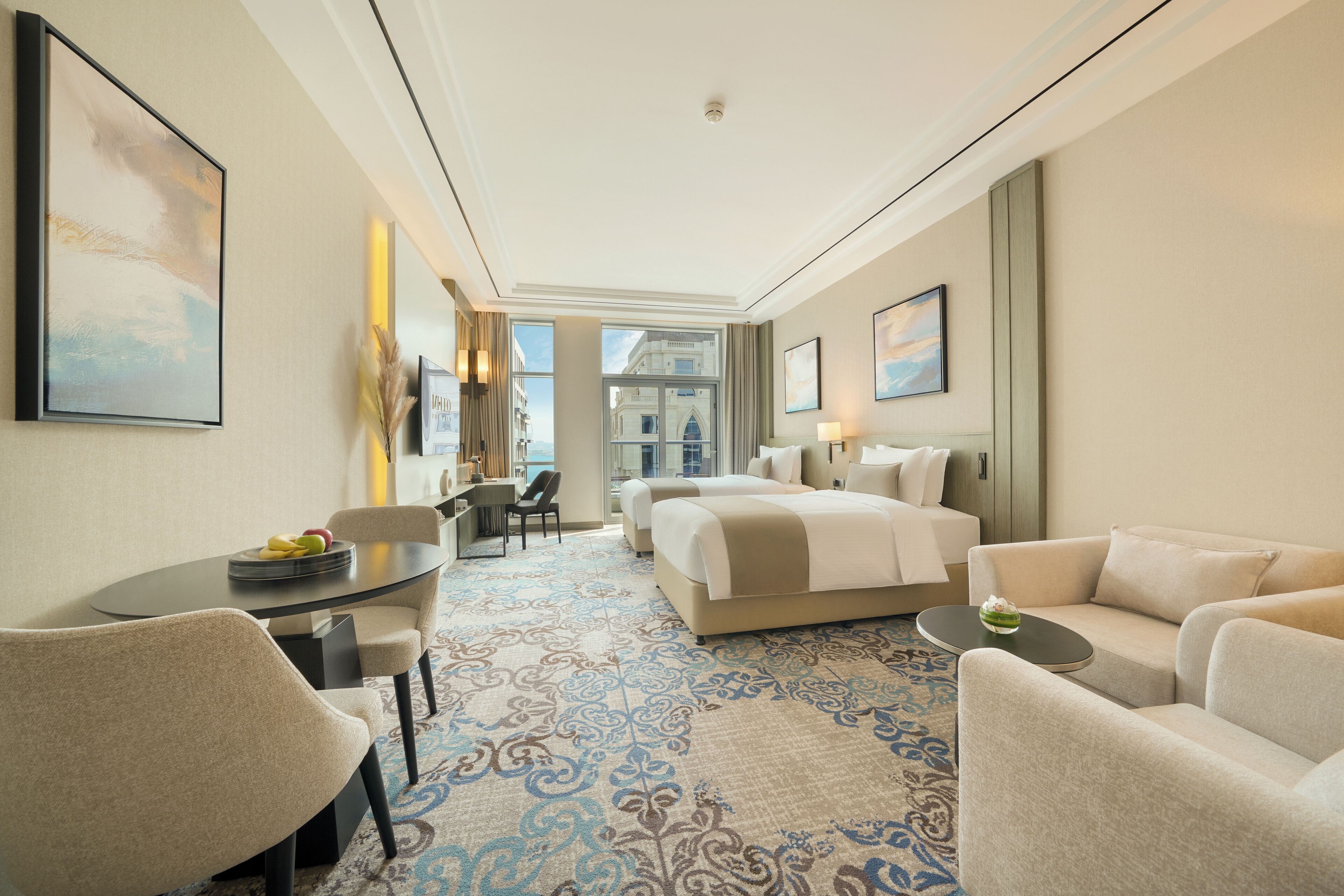 A hotel room with a bed, a sofa, a desk, and a chair. There is a window with a view of buildings.
