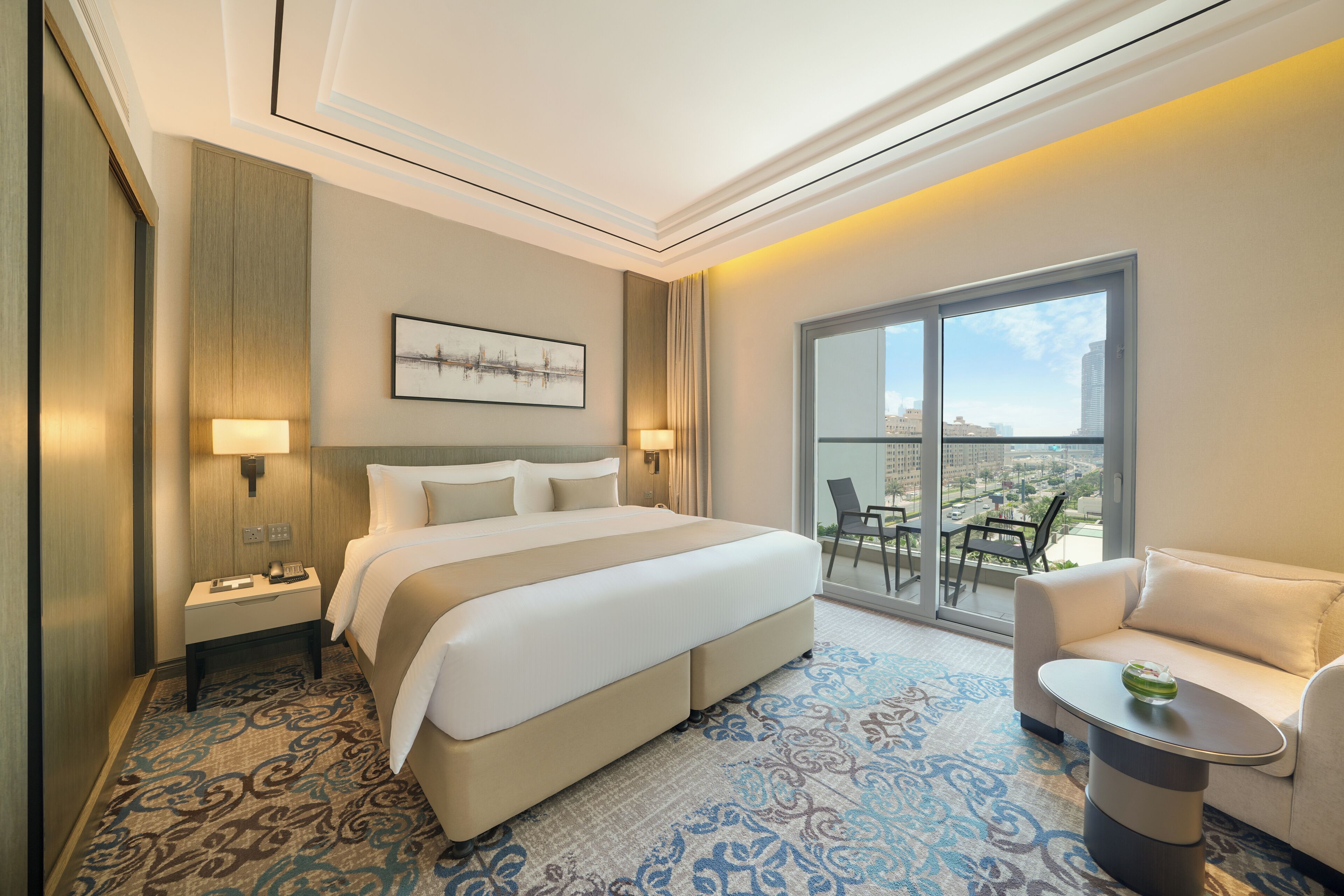 A hotel room with a large bed, a sofa, a small table, and a balcony with a view of the city.