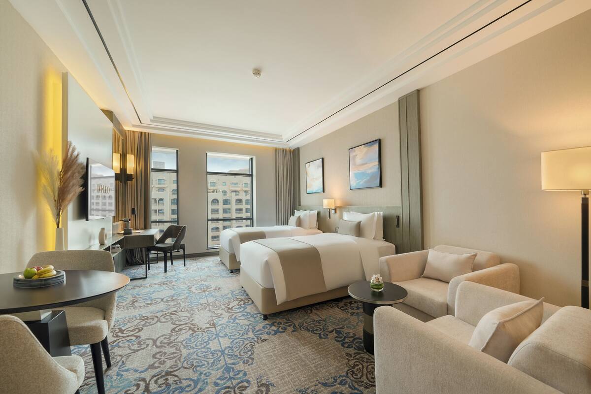 A hotel room with a large bed, two armchairs, a small table, and a desk. There is a window with a view of buildings outside.