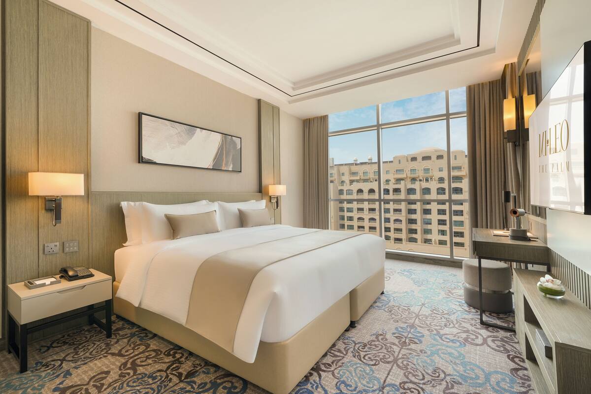 A hotel room with a large bed, a desk, a chair, a lamp, a phone, and a view of the city through large windows.