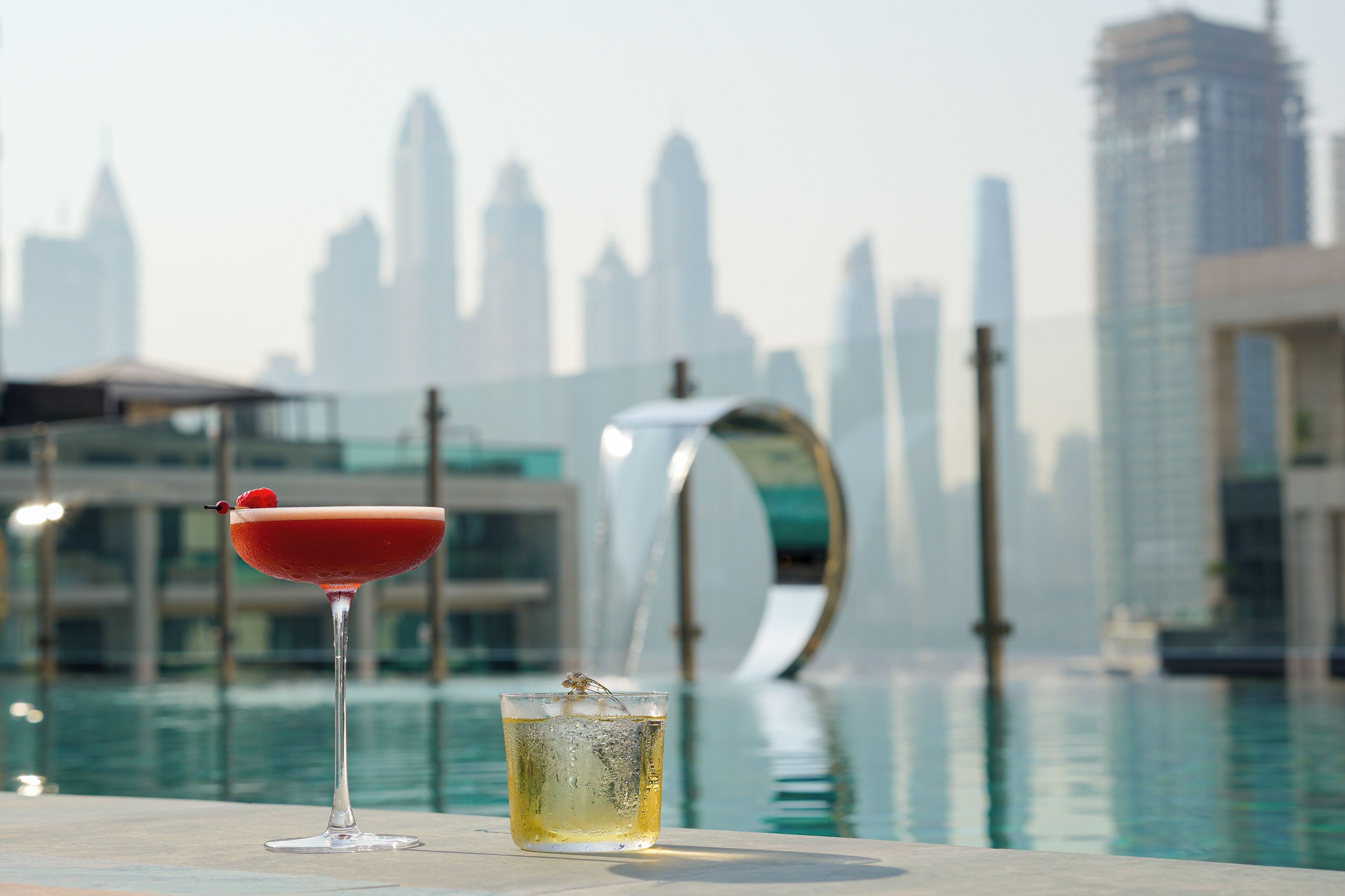 A cocktail glass with a red rim and a cherry on a table by a poolside with a city skyline in the background.