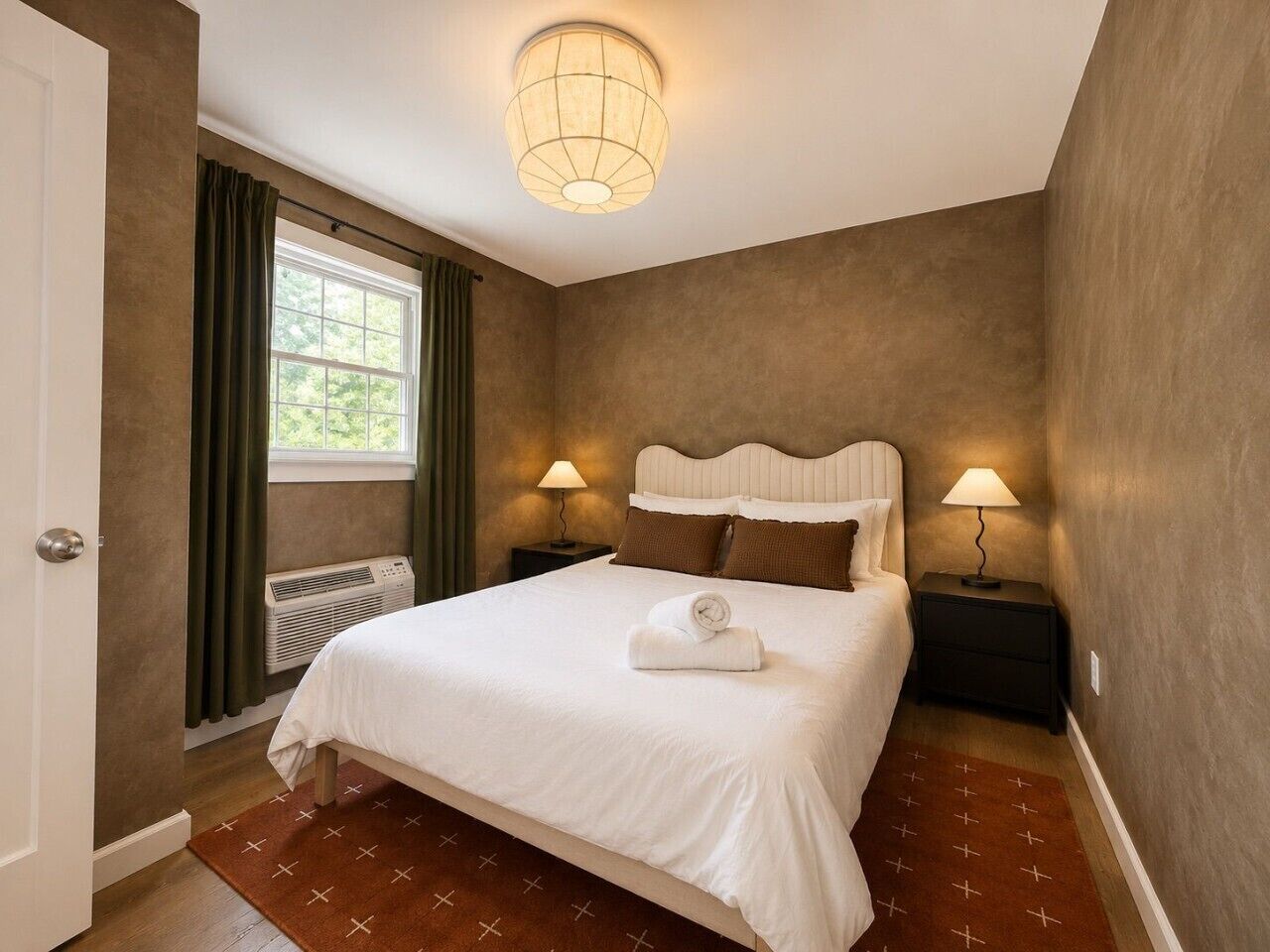 Une chambre à coucher comprenant un lit, deux lampes de chevet, une fenêtre avec des rideaux, un radiateur et un luminaire au plafond.