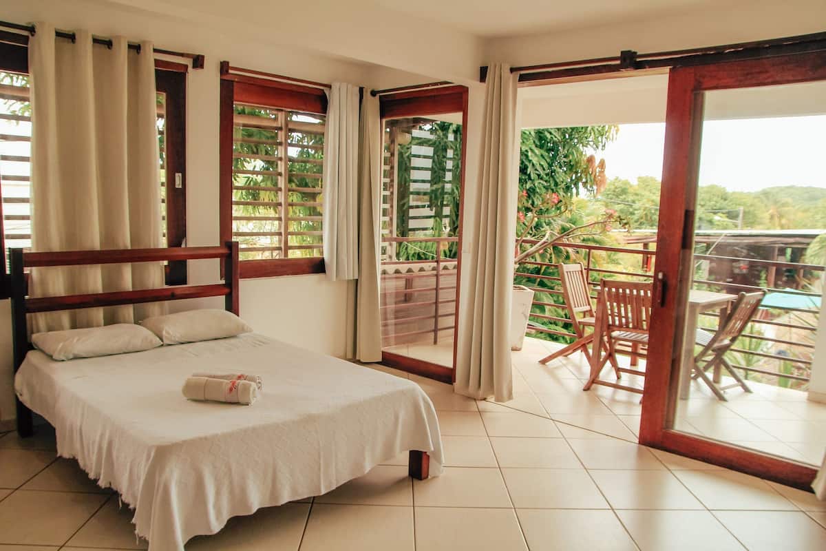 A hotel room with a bed, a balcony with a table and chairs, and a view of greenery.