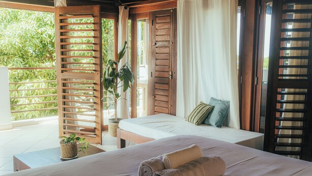 Un dormitorio con cama, almohadas, una toalla enrollada, una planta en maceta y una ventana con vistas a la vegetación.