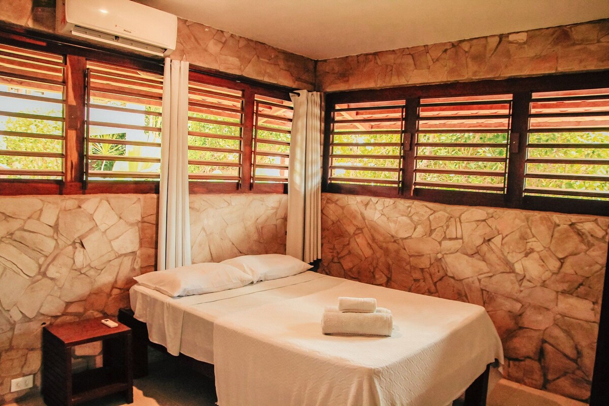 A bedroom with a stone wall, a bed with white linens, a bedside table, and a window with wooden shutters.