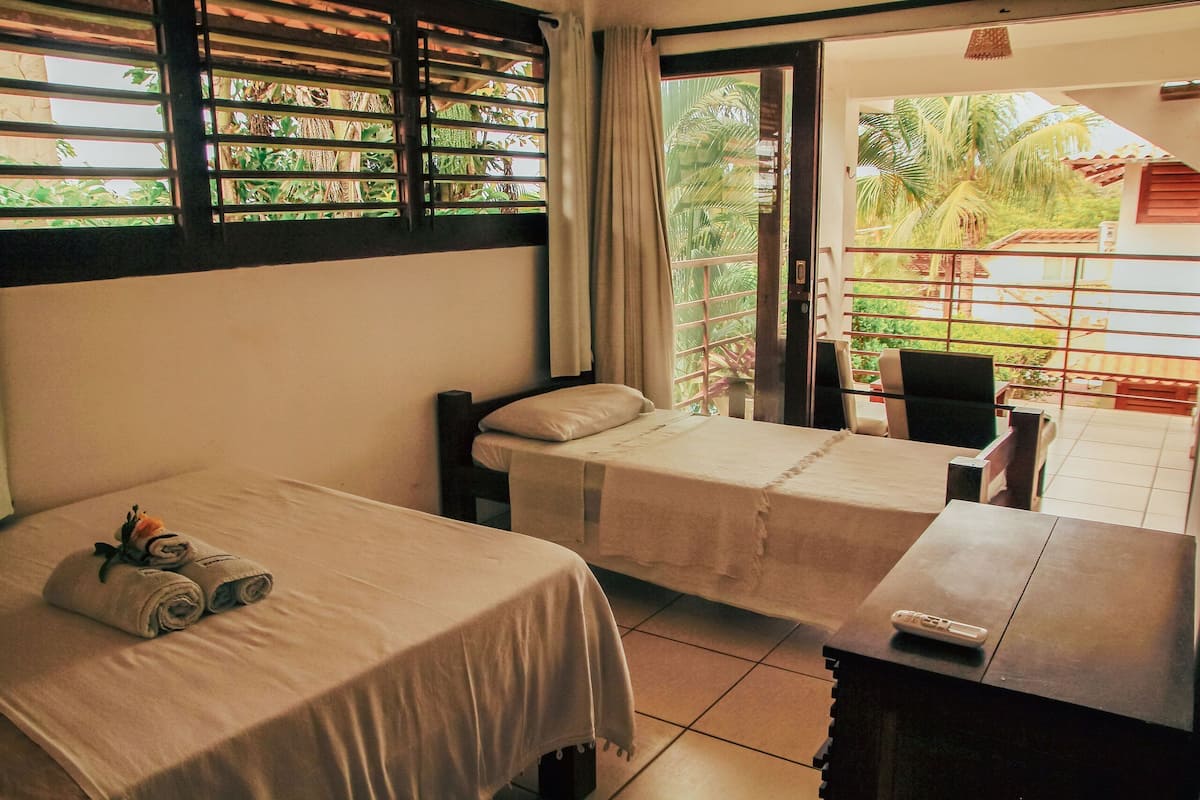 A hotel room with two beds, a balcony with a view of palm trees, and a table with a remote control.
