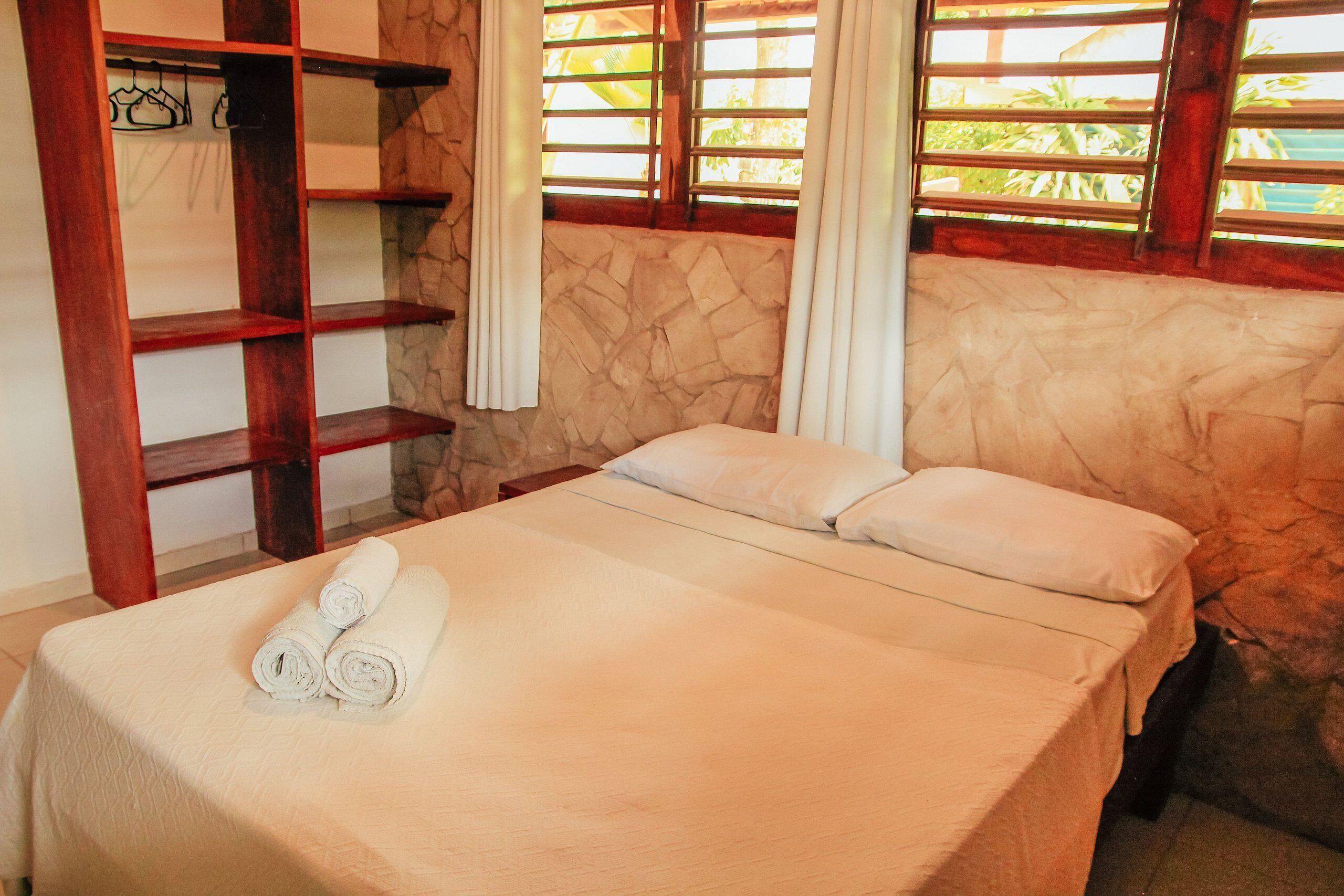 A neatly made bed with rolled towels, a wooden shelf unit, and a stone-textured wall.
