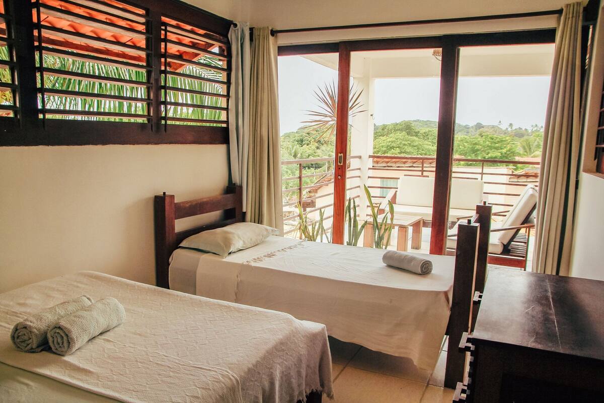 A room with two beds, a wooden headboard, a desk, and a balcony with a view of greenery.