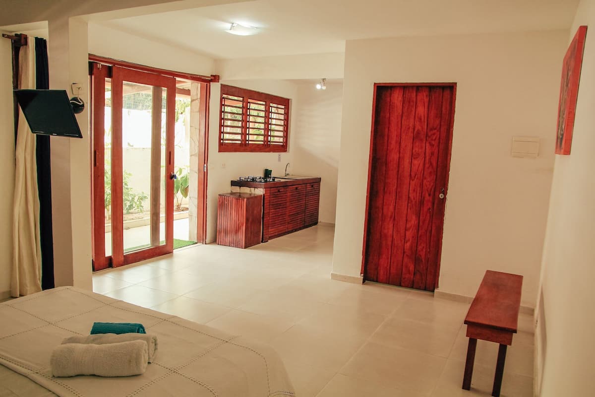 A hotel room with a bed, a small table, a kitchen area with a sink, and a sliding glass door leading to an outdoor area.