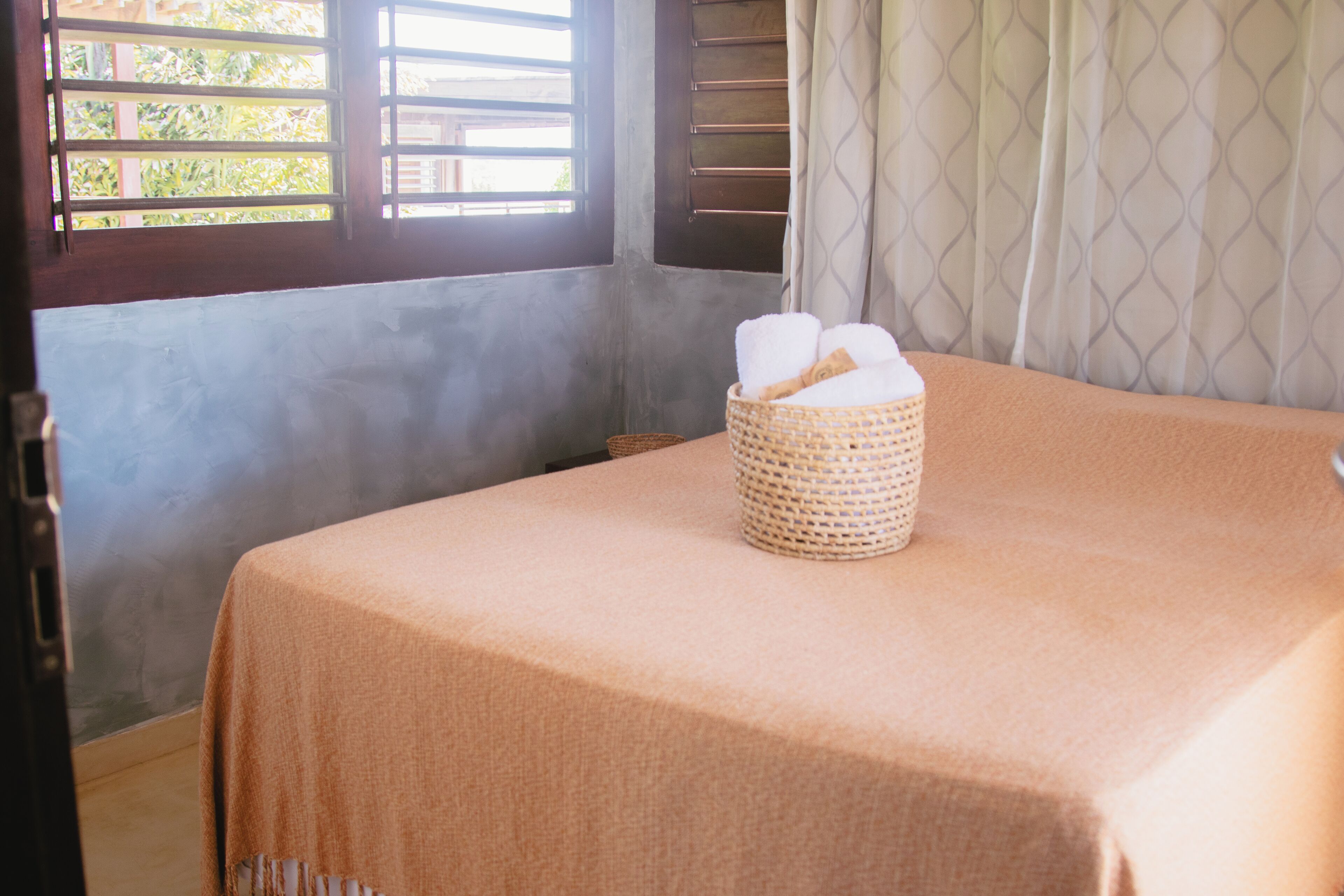 Une chambre avec un lit recouvert d’une couverture beige, un panier en osier contenant des serviettes posé sur le lit, une fenêtre avec des volets en bois et un rideau à motifs.