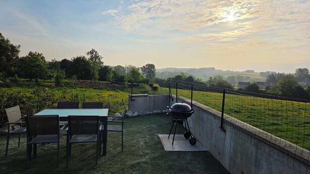 Een terras buiten met een tafel en stoelen, een barbecue en uitzicht op een groen veld en gebouwen in de verte.