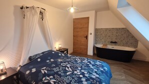 Een slaapkamer met een bed, nachtkastje, lamp, en een badkamer met bad en douche.