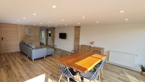 Een moderne woonkamer met een houten vloer, een grijze bank, een houten eettafel met stoelen en een metselwerk muur met een klok.