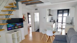 A modern living space with a flat-screen TV, a dining area with a glass table and white chairs, a grey sofa, and a kitchen area with a sink and stove.