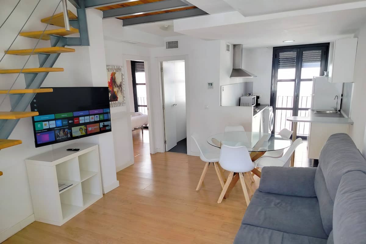 A modern living space with a flat-screen TV, a dining area with a glass table and white chairs, a grey sofa, and a kitchen area with a sink and stove.