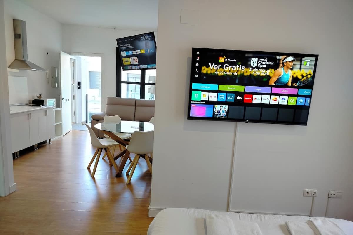A modern hotel room with a large flat-screen TV mounted on the wall displaying various channels. There is a dining area with a round table and chairs, and a kitchenette in the background.