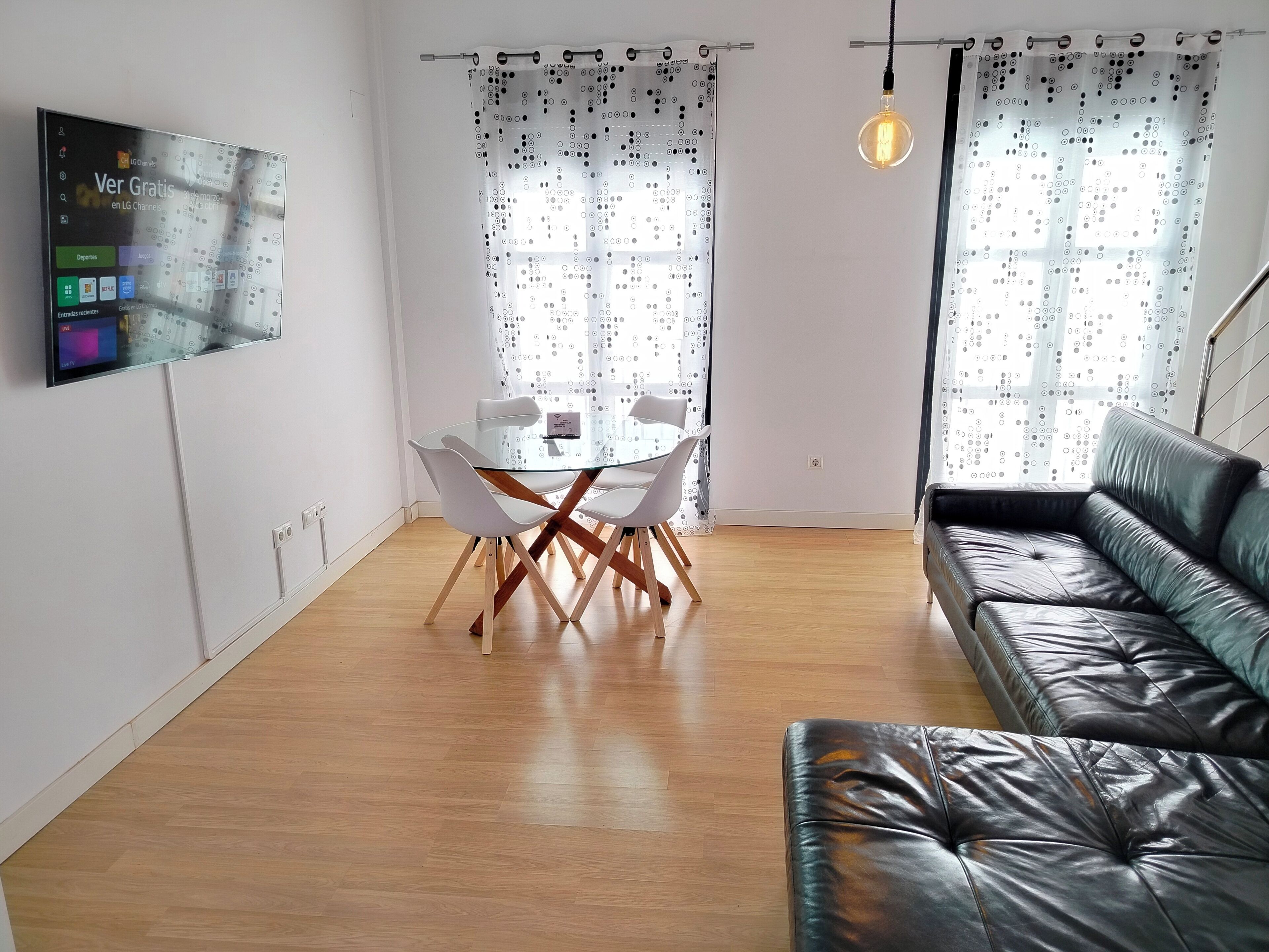 A modern living room with a black leather sofa, a glass dining table with white chairs, and a TV mounted on the wall.