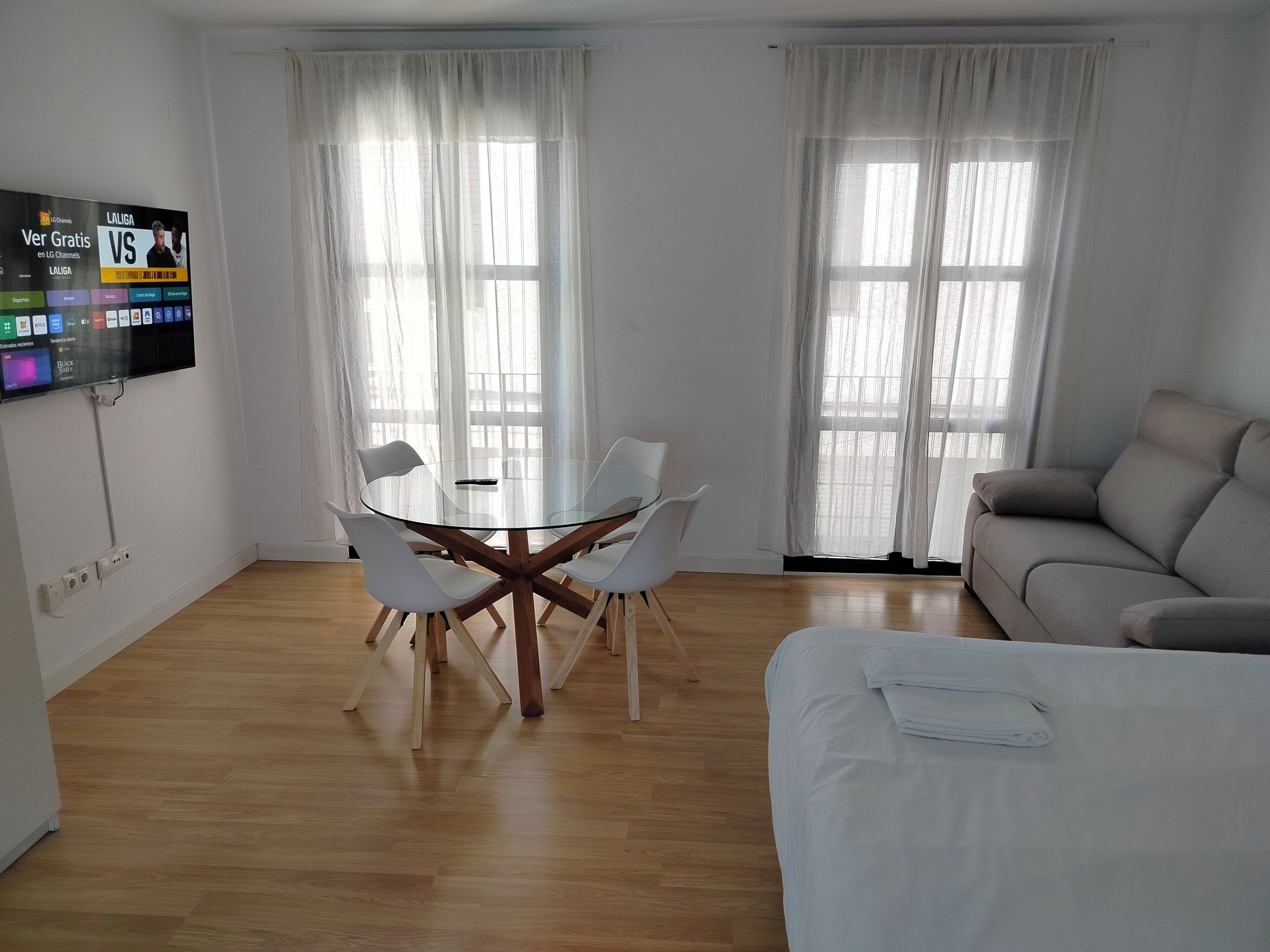 Een moderne woonkamer met een ronde glazen tafel, witte stoelen, een beige bank en een tv aan de muur waarop een sportzender wordt vertoond.